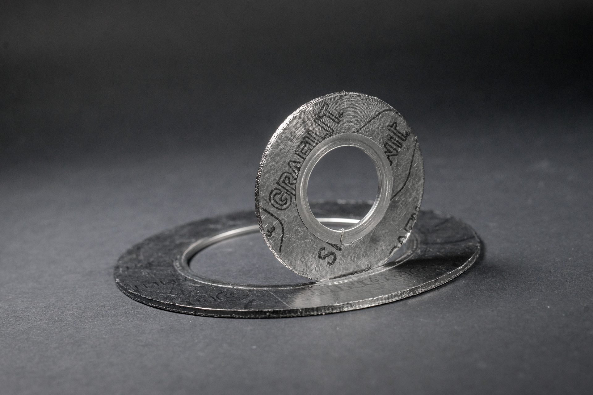 Metal technical gaskets of different sizes lying against a dark background, with one threaded through another.
