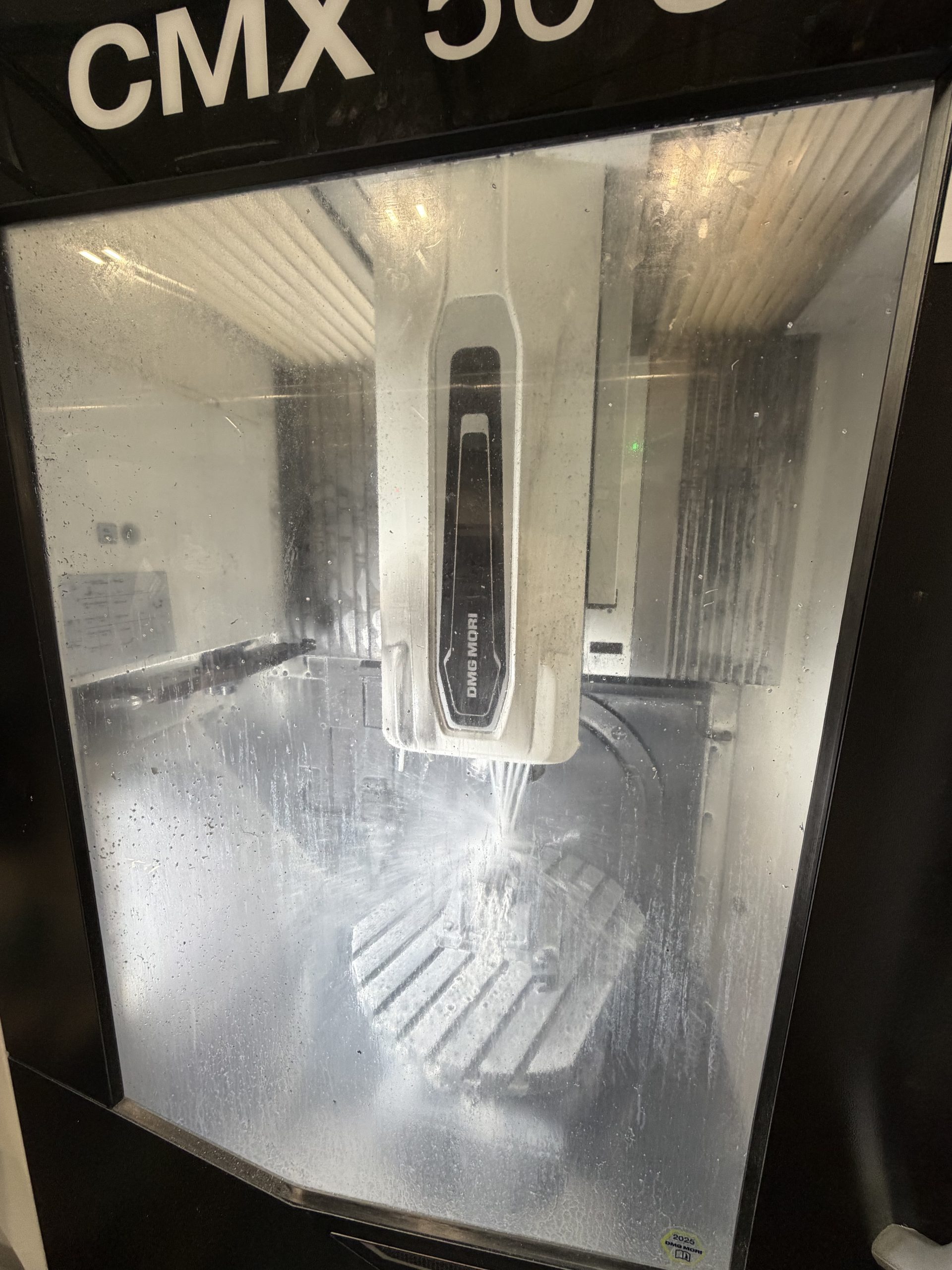 A modern CNC milling machine behind a closed window with water droplets, operating in a production plant.
