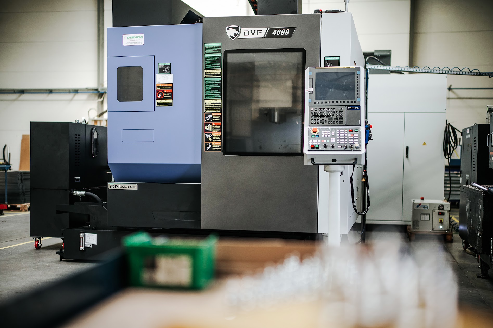 A modern CNC machine in a production hall, with a glazed working chamber and a control panel on a stand.