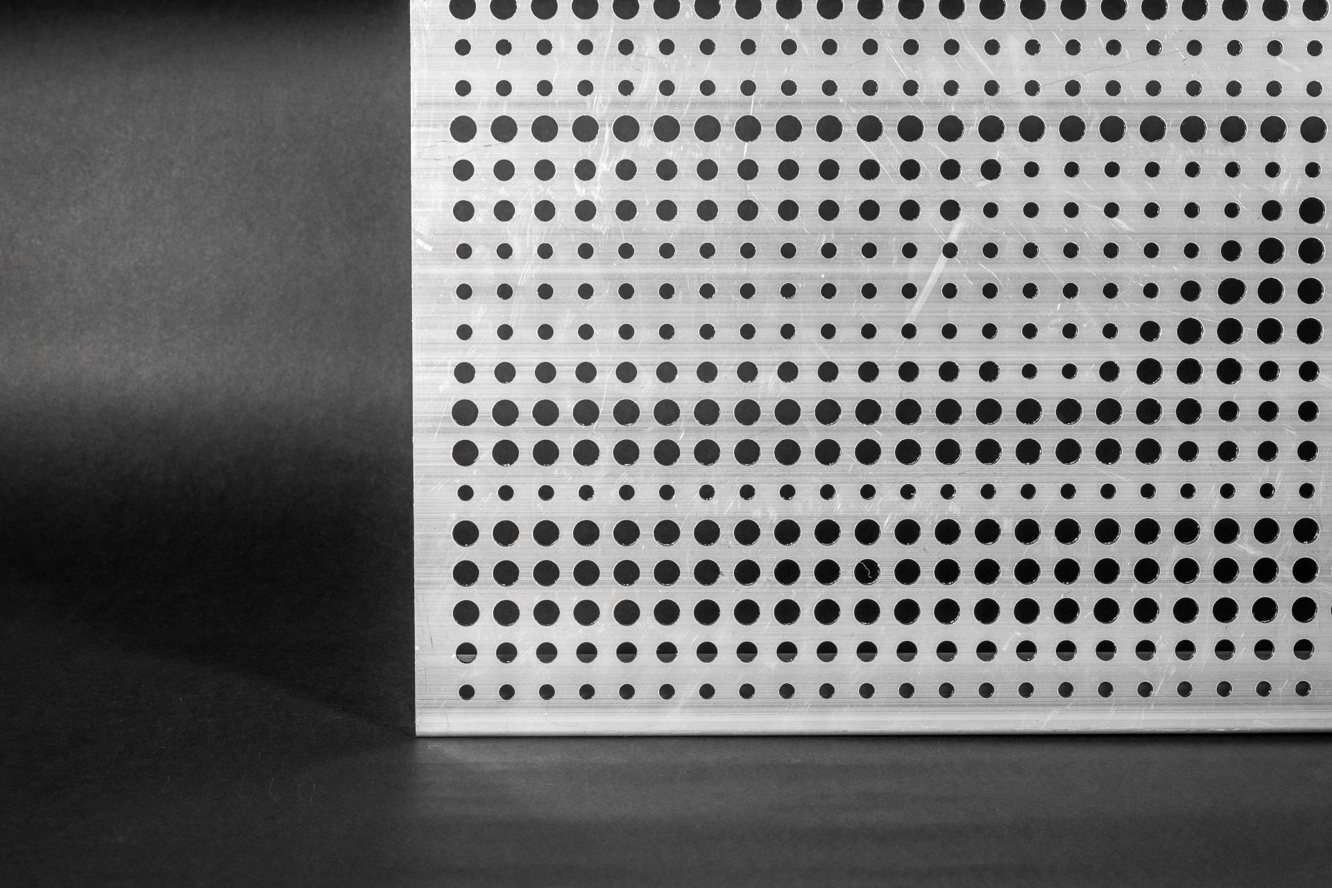 Perforated metal panel with rows of round holes of various diameters, set against a dark background.