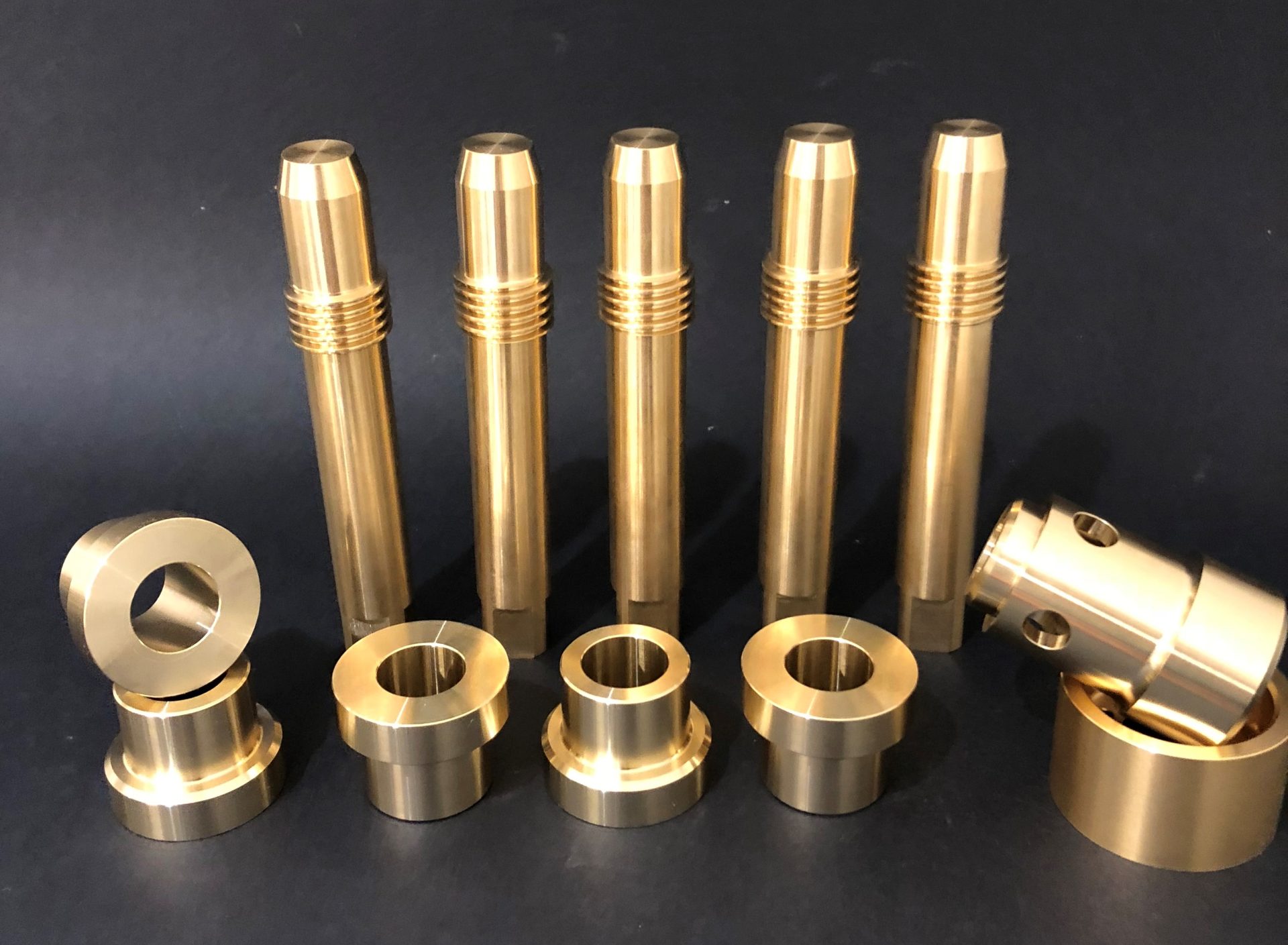 Five tall golden cylinders with threads on top and rings lying on a dark background next to a part with holes.