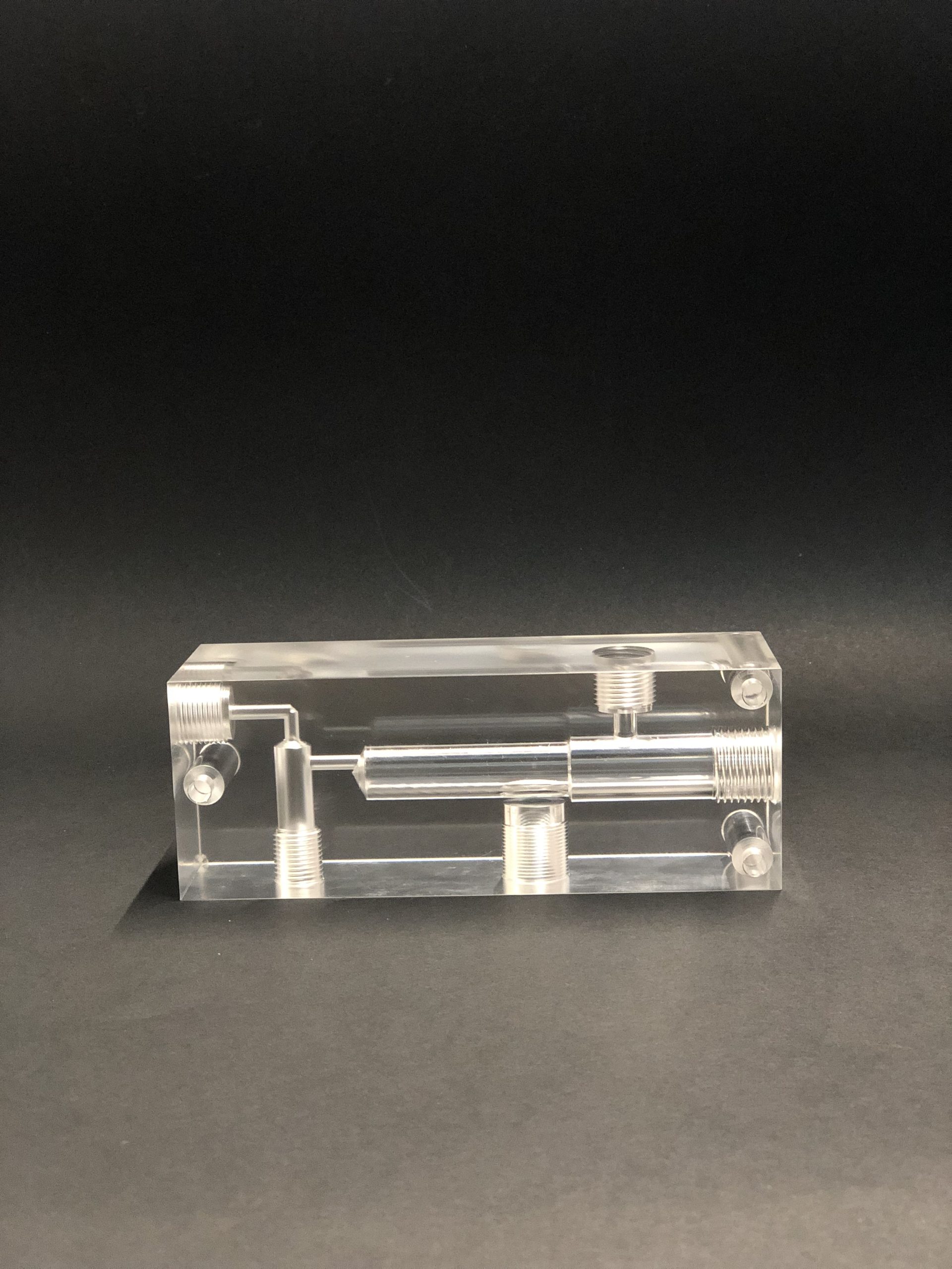A transparent rectangular block with internal channels, threaded holes and four screws at the corners on a dark table.