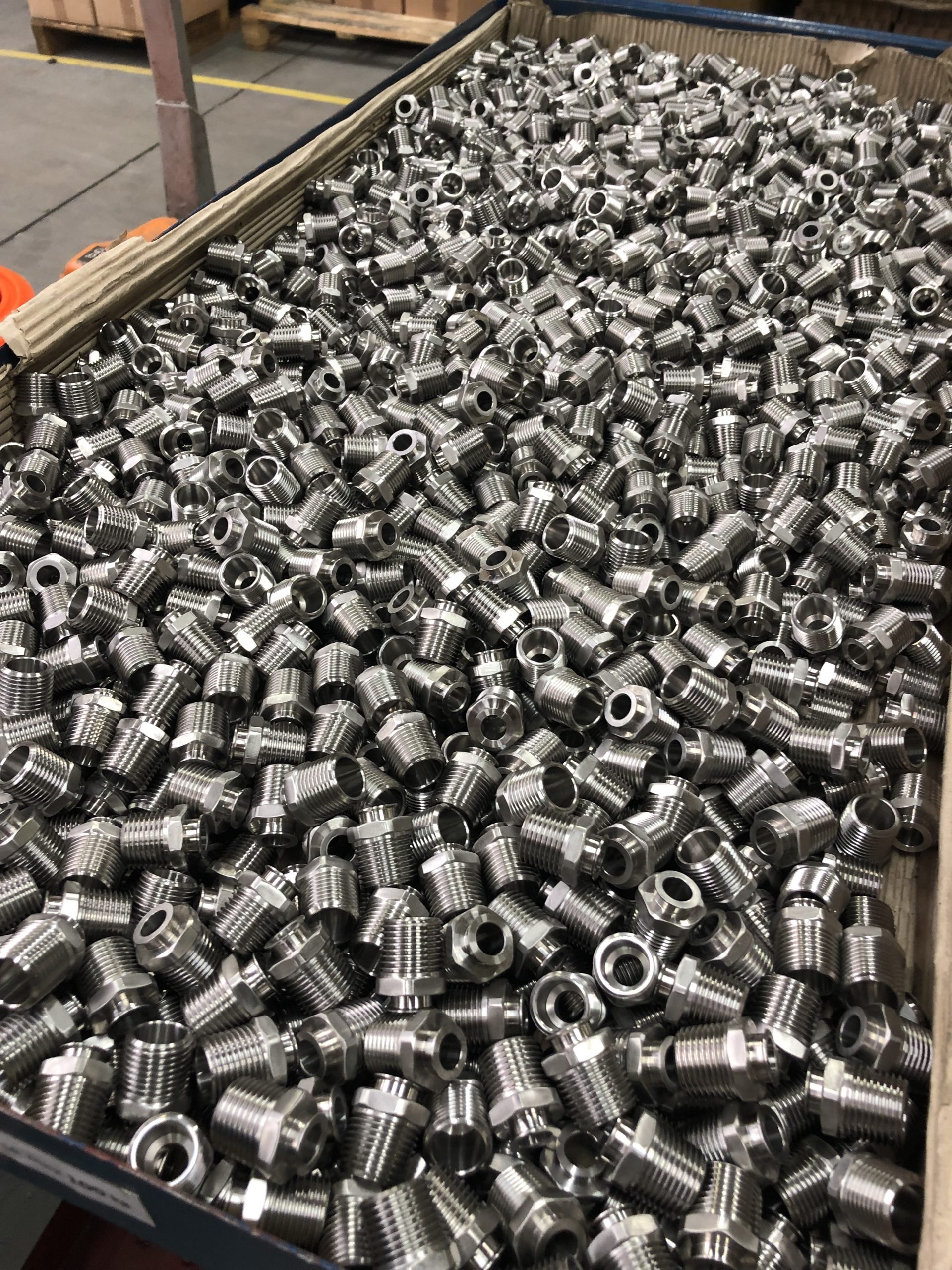 Shiny silver metal parts with an external thread fill a cardboard storage container to the brim.