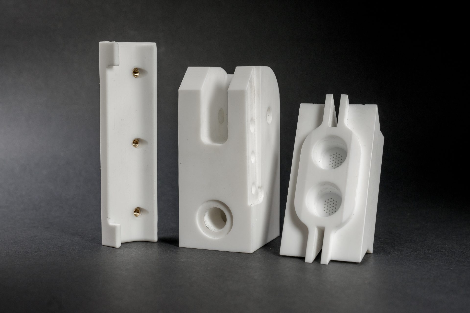 Three white plastic parts with holes, recesses and profiled edges against a dark studio background.