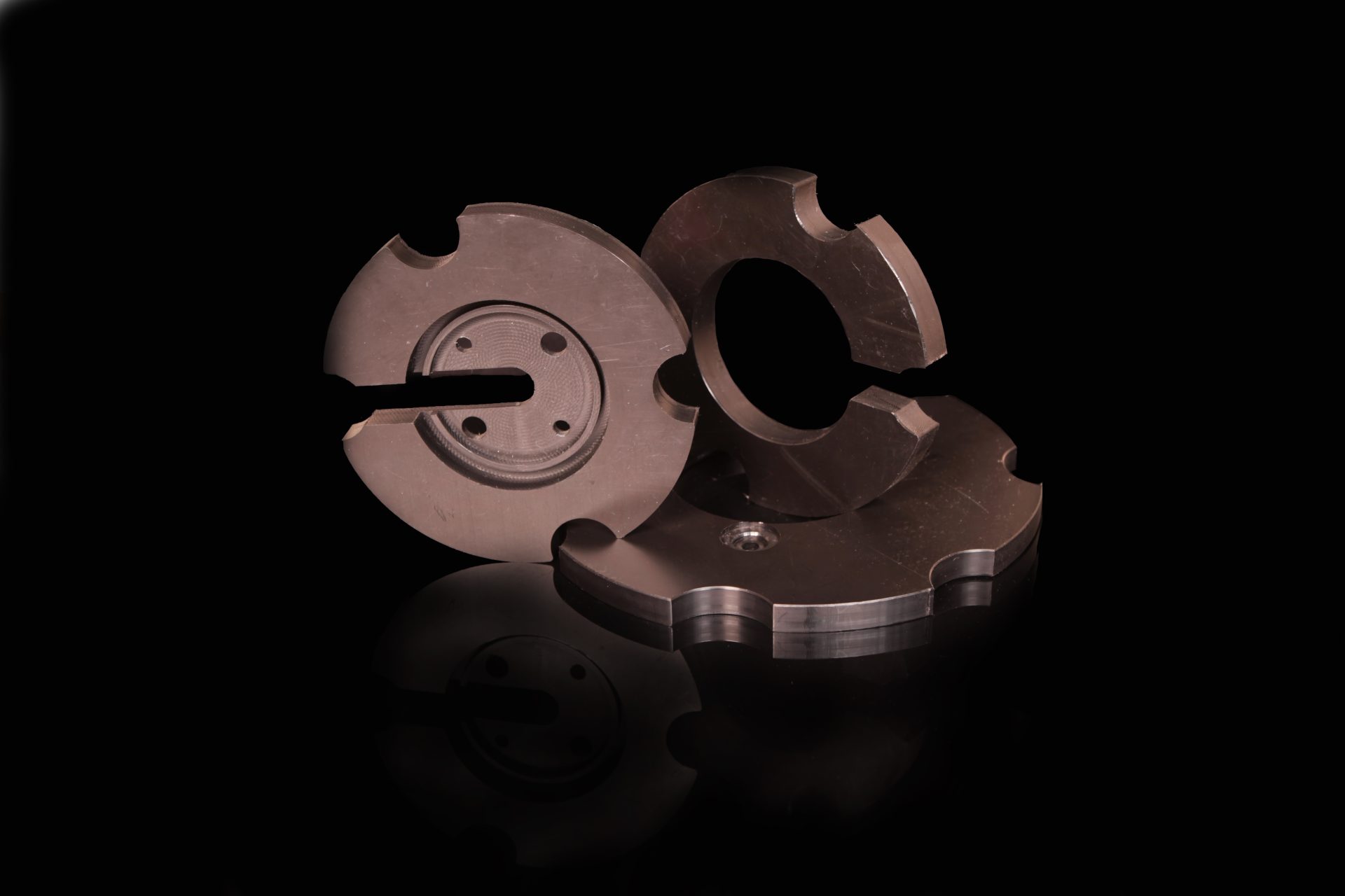 Three dark grey circular parts with holes and cut-outs lying on a black surface with a reflection.