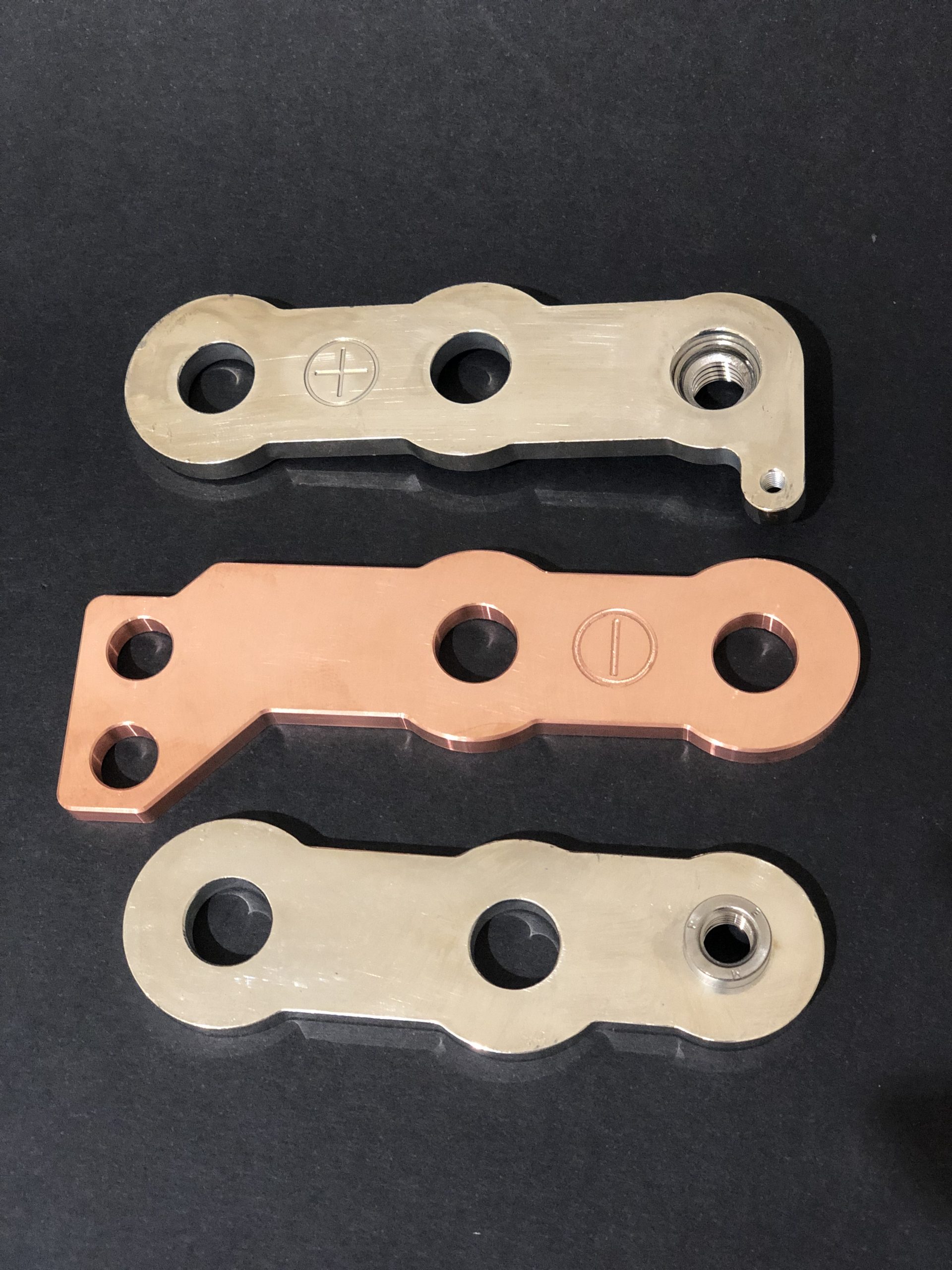 Three metal parts arranged on a black background: two silver ones with threaded holes and one copper.